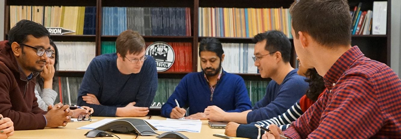 group of eight students sitting around a table and looking at a document, bookshelves are lined along the wall behind them