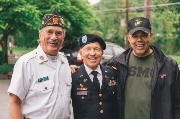 three generations of veterans stand side by side smiling at the camera, trees are visible behind them